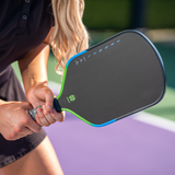 A close-up lifestyle shot of a person holding a pickleball paddle on a purple and green pickleball court. The paddle has a dark gray face, a bright blue and green frame, and the brand name 'THE DAY' is visible on the paddle.
